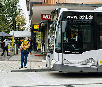 Eine Pendlerin steht an einer Bushaltestelle vor einem wartenden Bus.