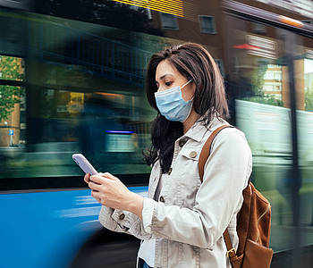 Frau mit Maske steht vor einem Bus und schaut auf ihr Smartphone