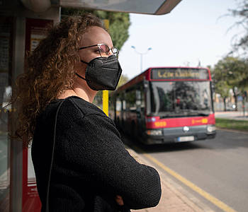 Frau mit FFP2-Maske steht an einer Bus-Haltestelle